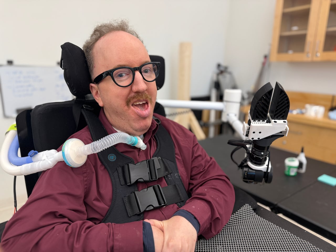 A man with glasses and a burgundy shirt sits in a wheelchair with a black and white robot claw attached to it.