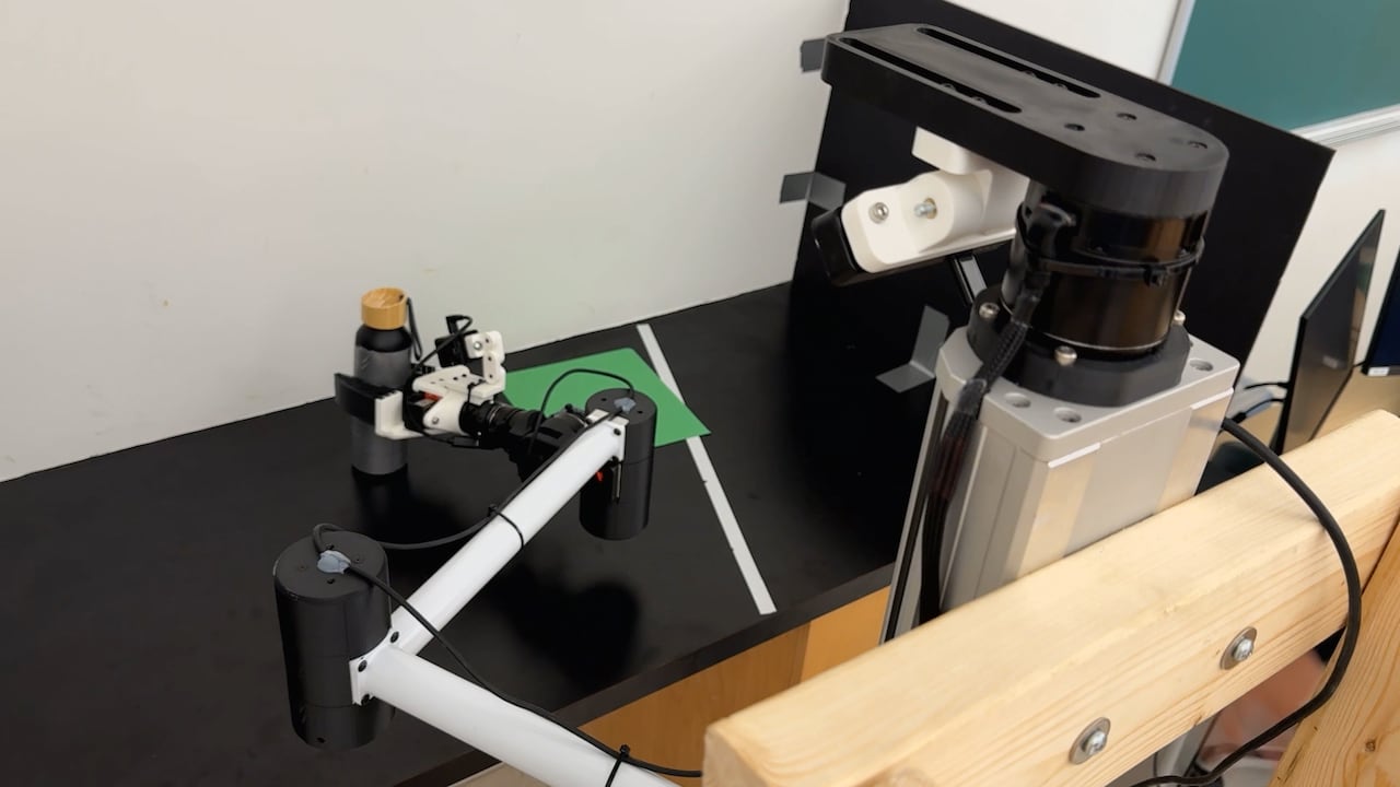 A white robotic arm reaching for a water bottle. A camera on a wooden post is in the foreground.