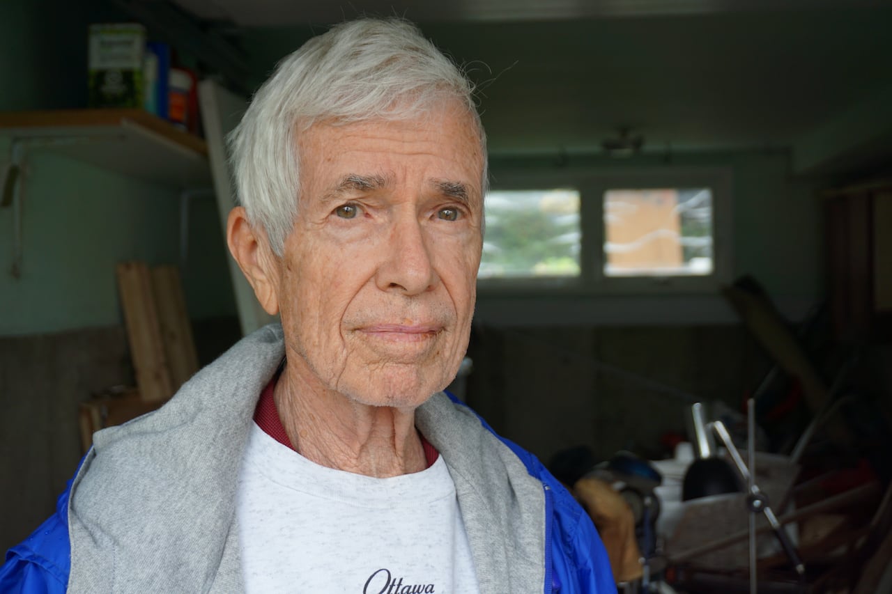 An elderly man with white hair and a blue jacket standing in a wrecked garage.