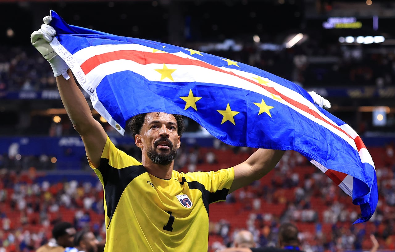A man in a soccer kit and goaltender gloves holds up the Cabo Verde flag in a stadium