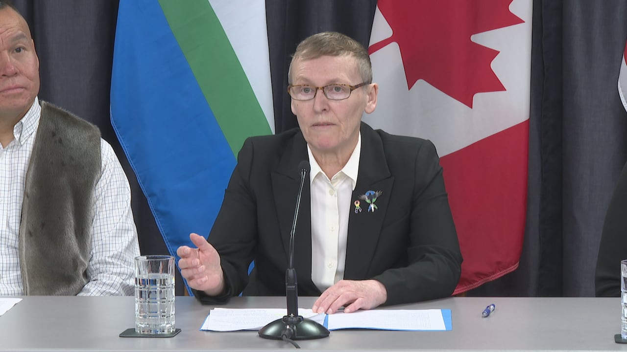 A woman in a suit jacket sits at a conference table and speaks into a microphone. To her right, also seated, is a man in a collared shirt and seal skin vest.