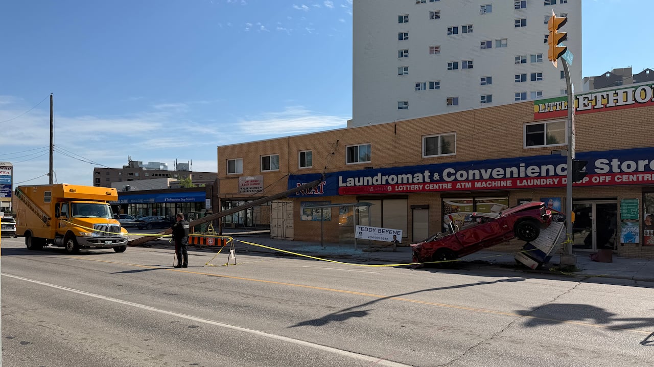 A city street is shown with a red vehicle crashed into a light post along a sidewalk. Yellow caution tape surrounds the scene