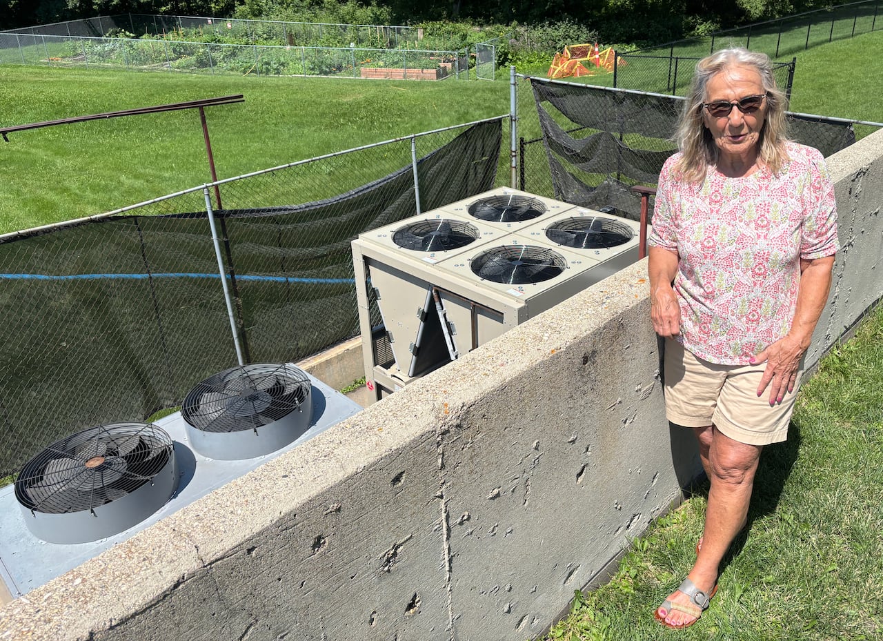 A woman with long grey hair stands on the grass, overlooking air conditioning units