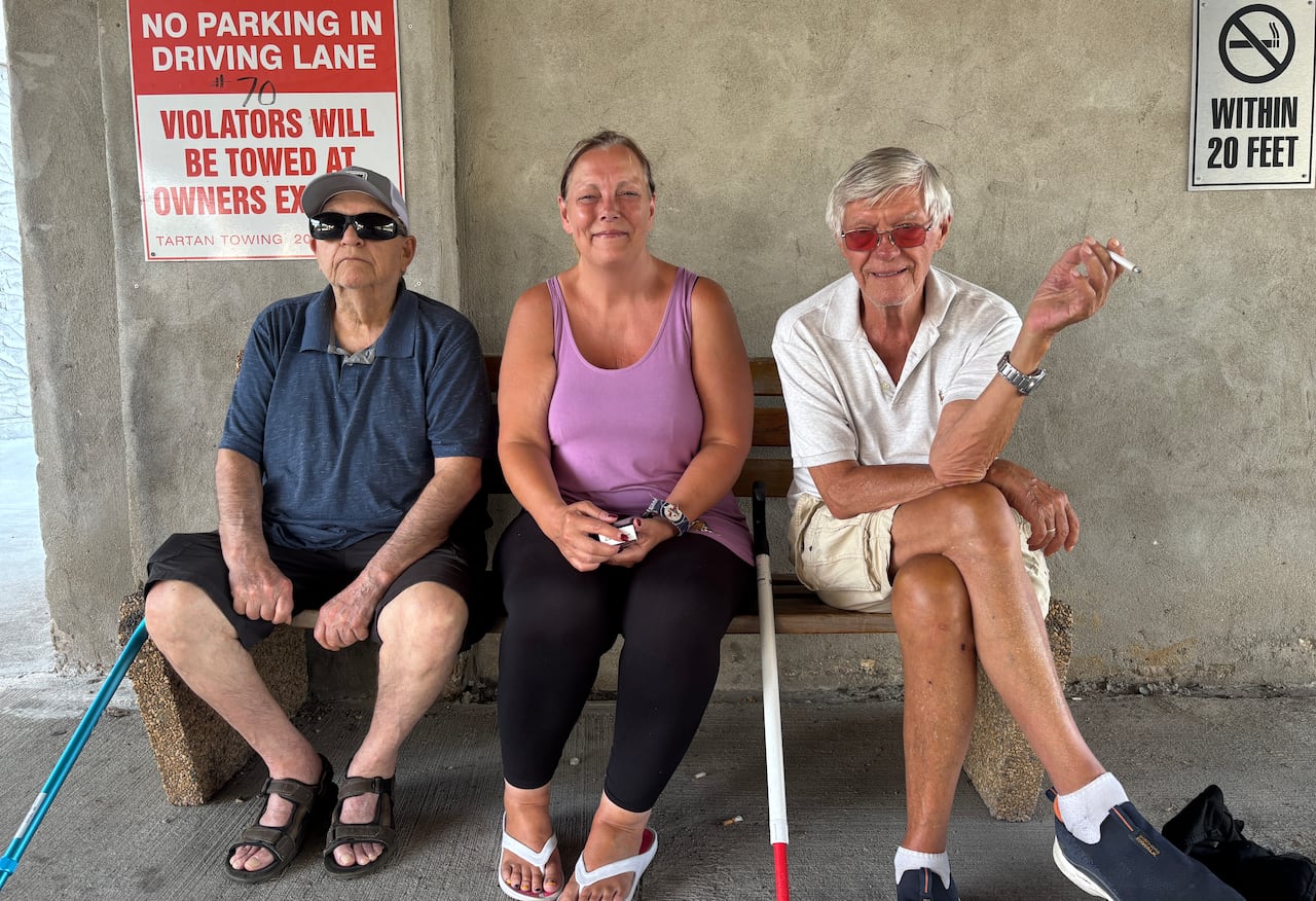 Three people sit on a bench set against a plain concrete wall with signs on it. Two canes rest against the bench and one person is holding a cigarette.