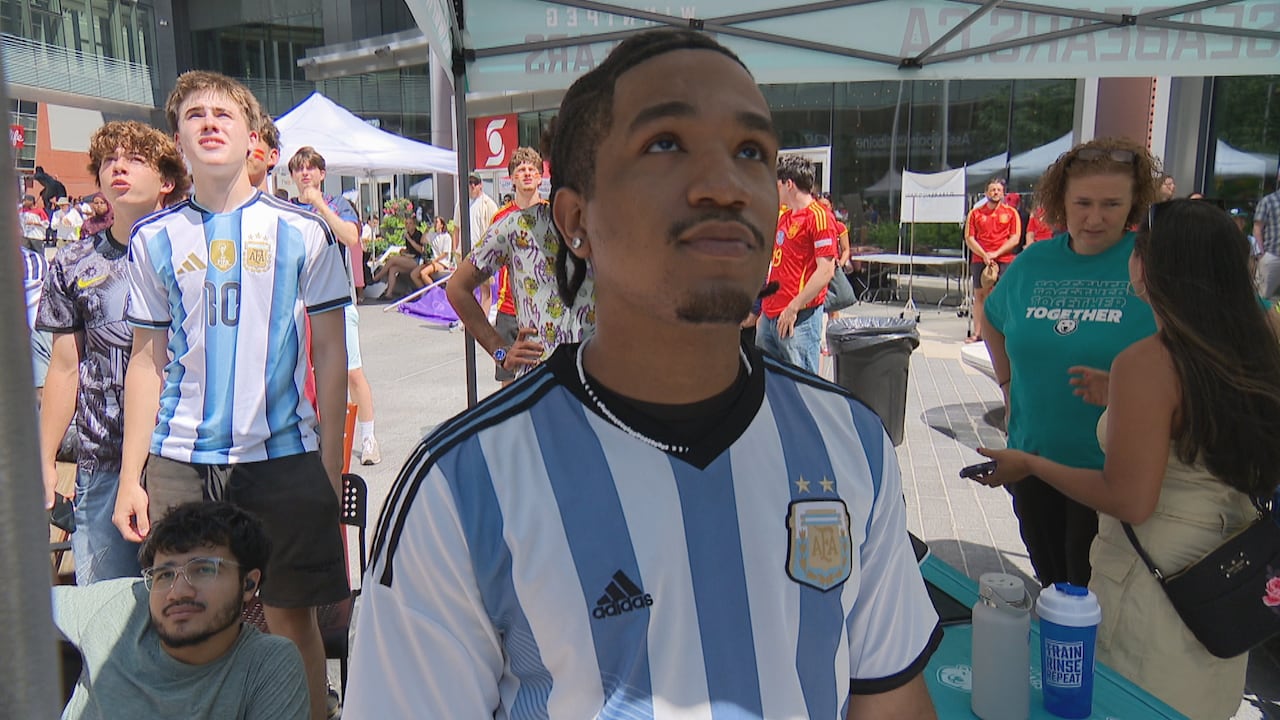 Fans in Argentinian soccer jerseys stand solemnly watching a screen.