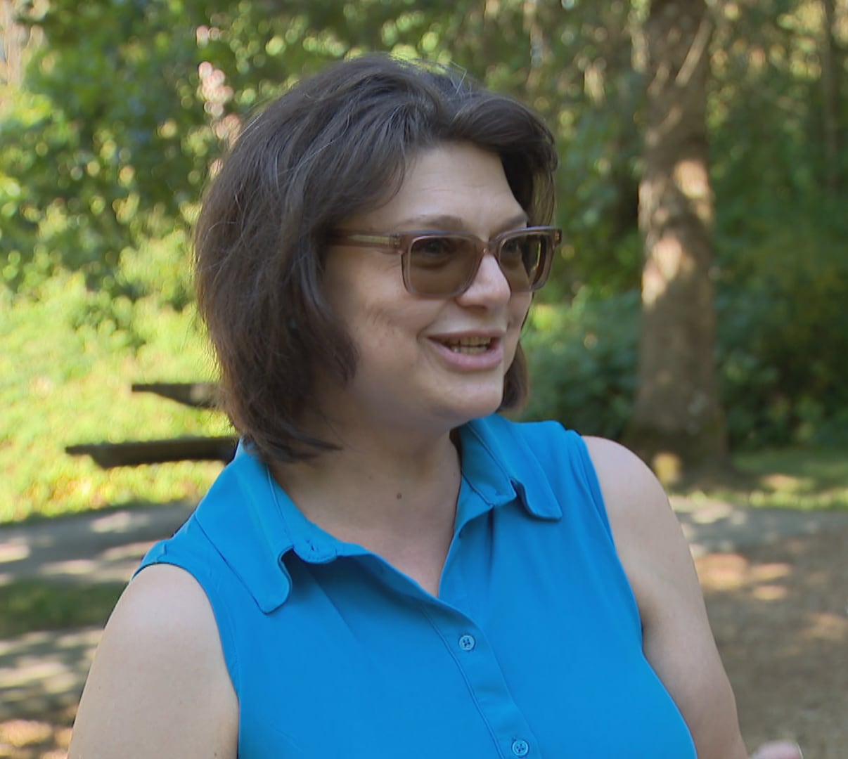 A woman with brown hair and sunglasses in a blue sleeveless top in a park.