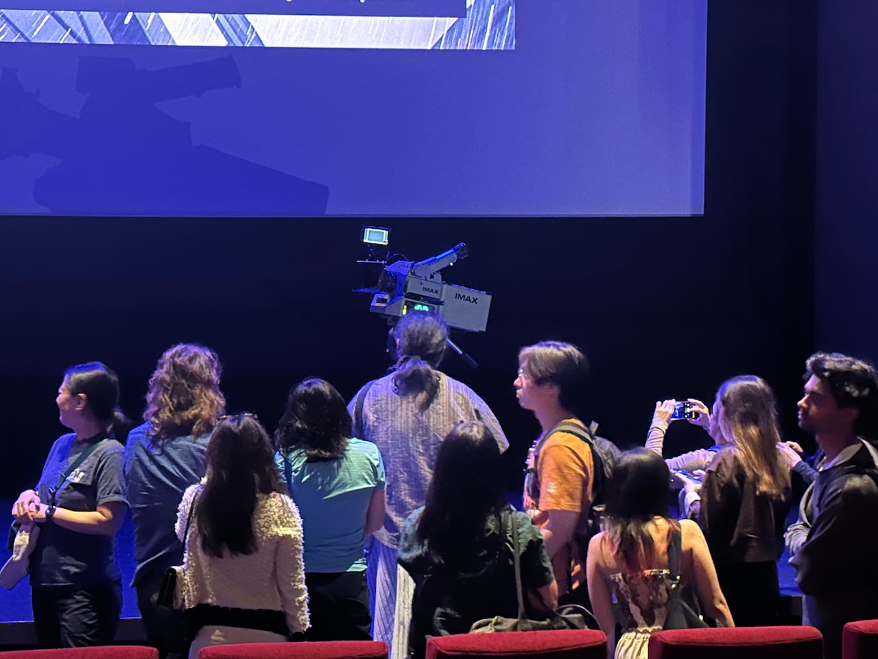 A video camera is seen on stage. People stand in front of it.