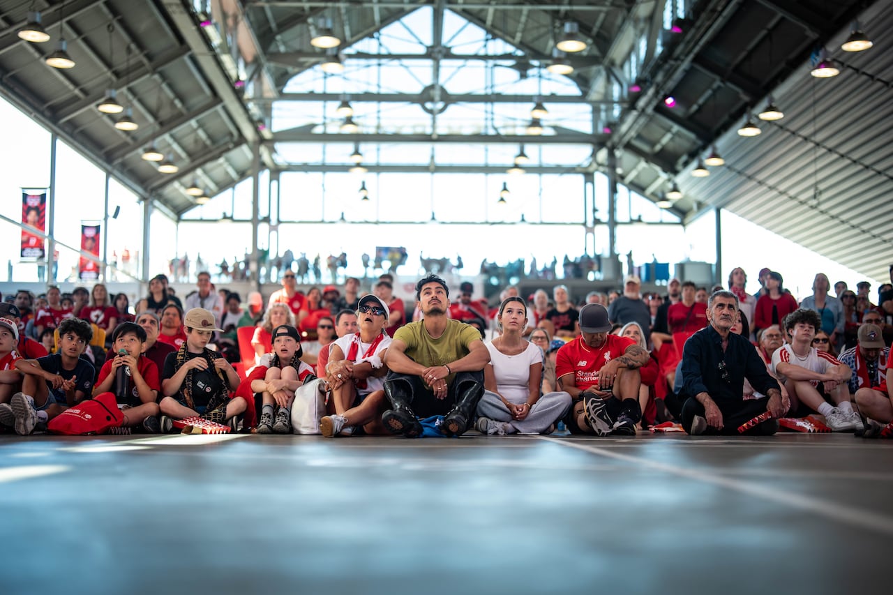 A group of people sit on the floor of a covered area, watching a game.