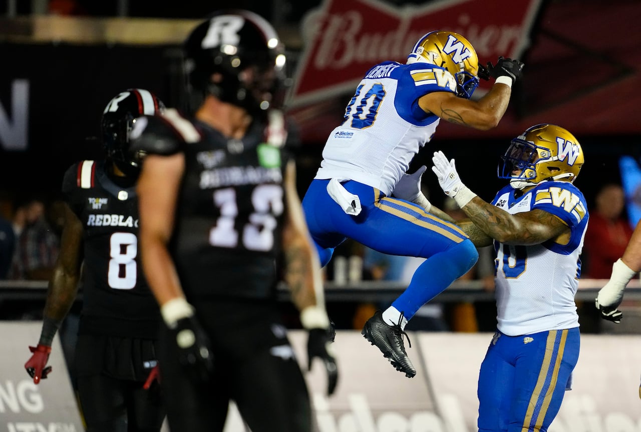 A football player in white and blue and gold jumps in the air in celebration while an opposing player in black and red looks on dejectedly.