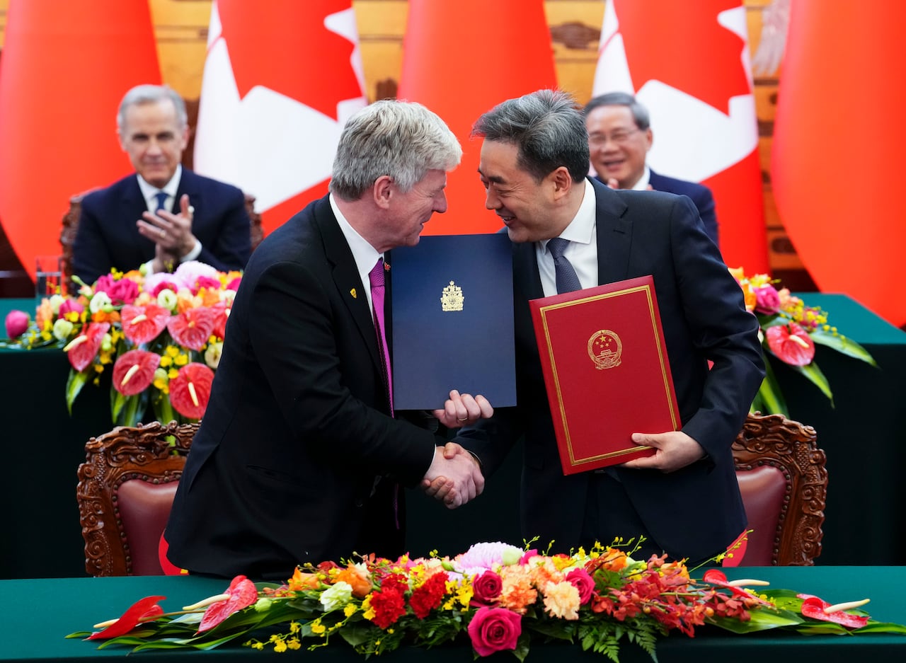 Two men holding folders shake hands in front of Chinese and Canadian flags.