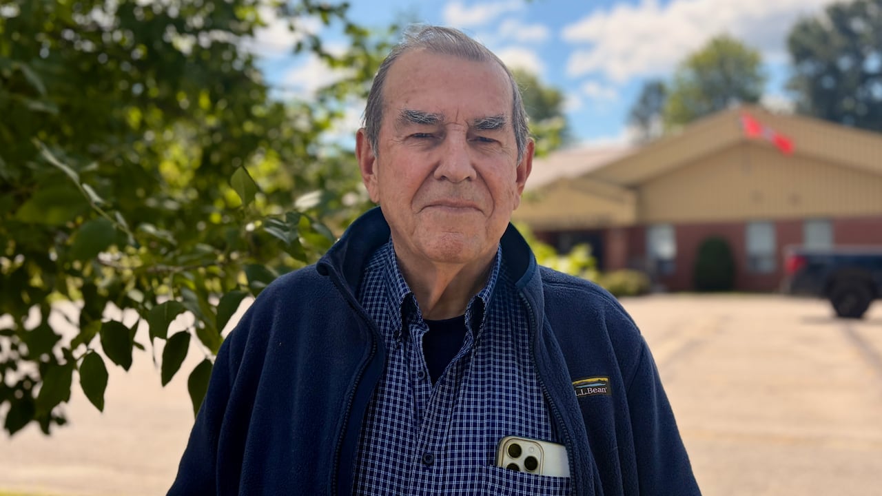 A man in a blue shirt and blue jacket poses for a photo outside on a sunny day.