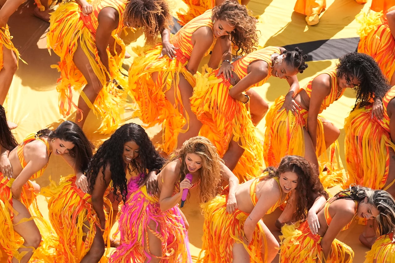 Shakira performing in a yellow and pink outfit with feathers, surrounded by other dancers