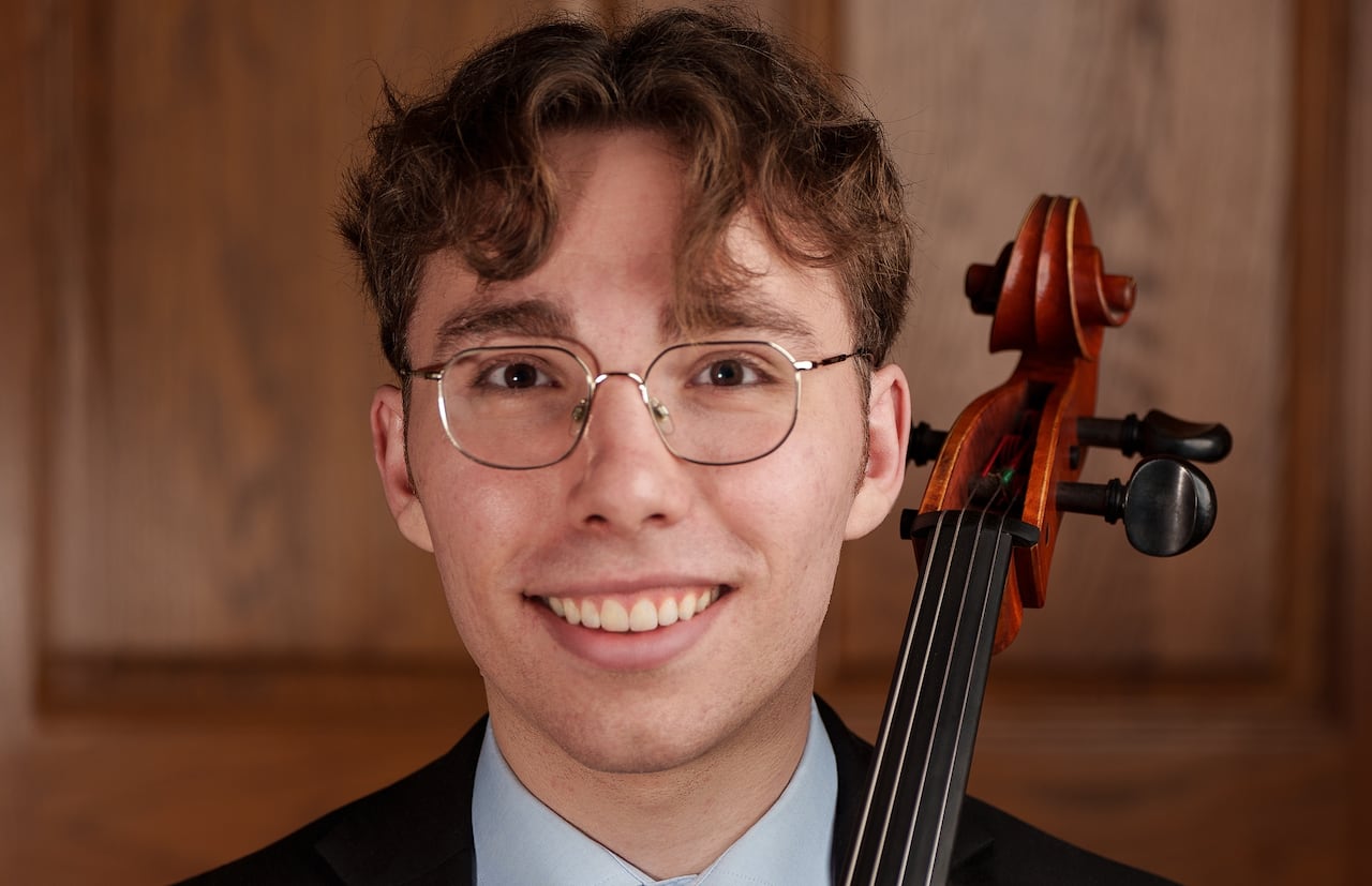 Elie Boissinot faces the camera with a smile on his face, holding his cello by the neck.