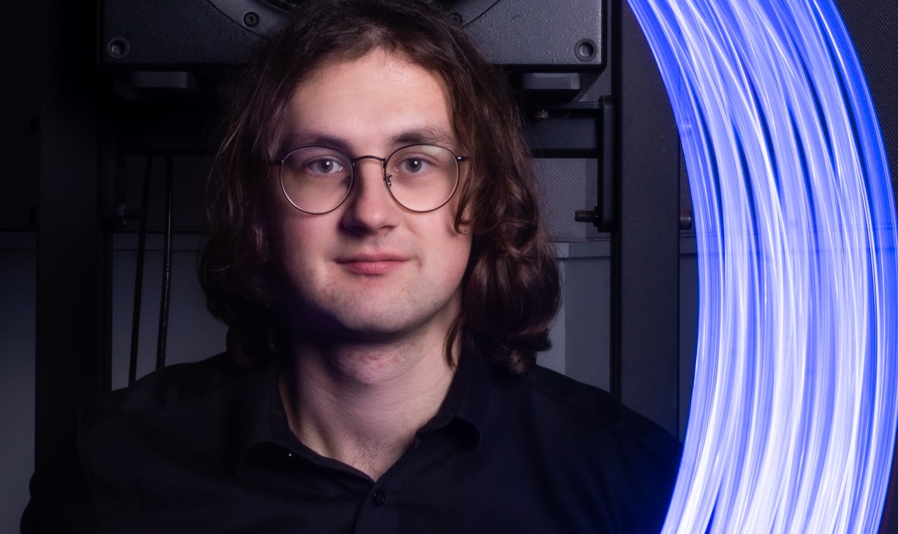 Dressed in black, Luke Blackmore faces the camera with a large speaker behind him and an arc of blue light floating in the space to his left.