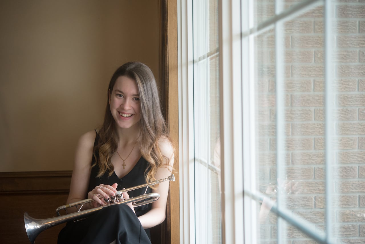Laura Kuri sits next to a window, holding her trumpet.