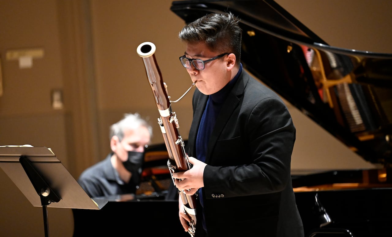 Bobby Thompson plays bassoon onstage, standing in front of a grand piano with the lid open.