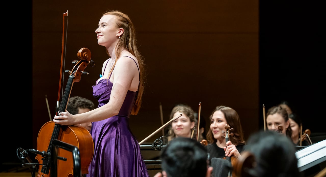 Wearing a purple gown, Sabina Sandvoss stands onstage in front of an orchestra while holding her cello.