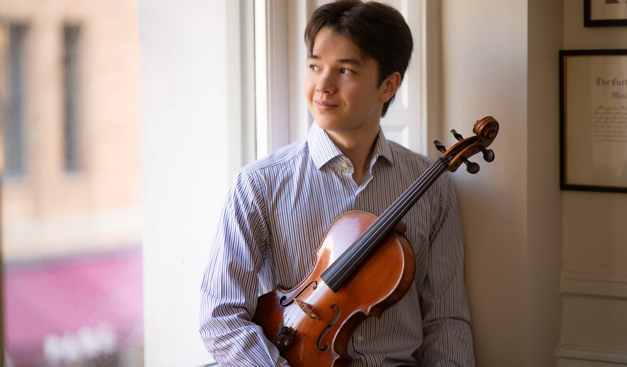 Davin Mar glances out a window while holding his viola under his arm.