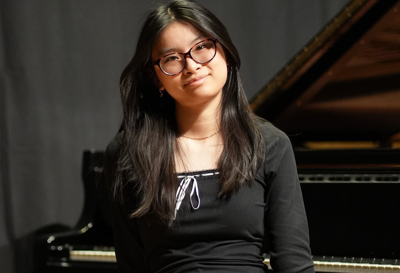Dressed in black, Angel Suet Kee Ng sits at the bench of a grand piano with the lid open.