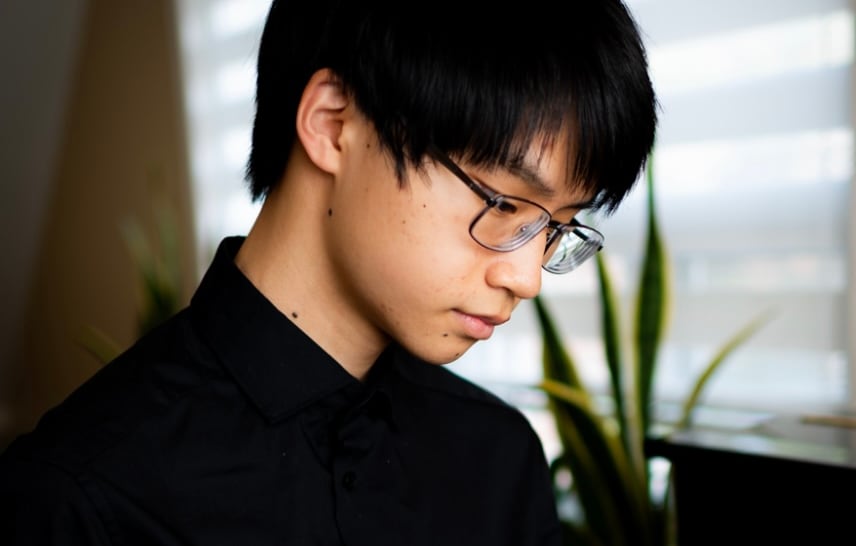 Wearing a black shirt, Kevin Wang looks wistfully away from the camera, standing in front of a window with the blind drawn.
