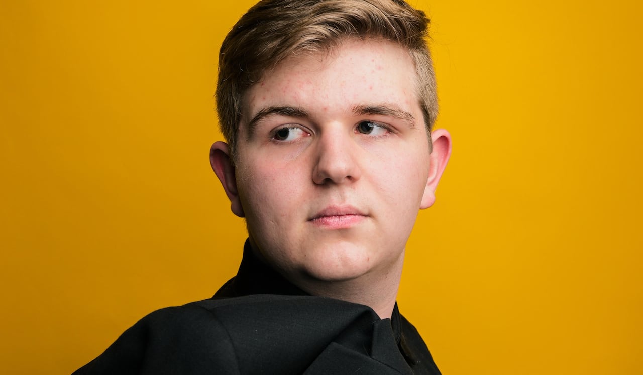 Against a yellow backdrop, Ashton Latimer looks away from the camera, with his black jacked tossed over his shoulder.