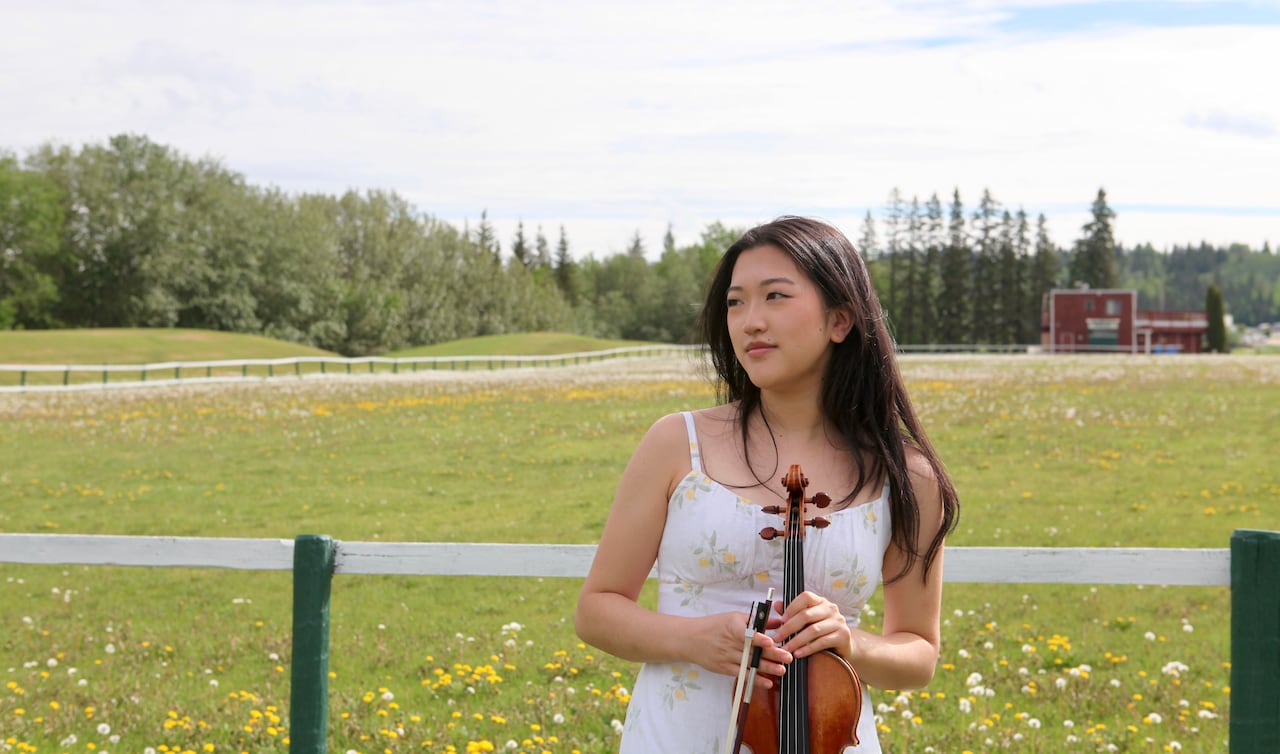 Holding her violin, Anna Yin stands in a lush, green field, looking off to the left.