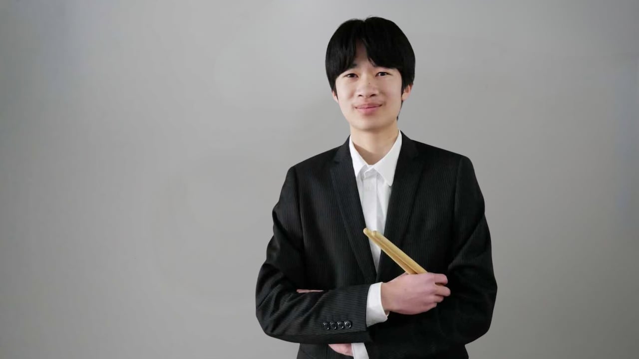 Ryan Gu, attired in black and white, holds a pair of drumsticks while smiling for the camera.