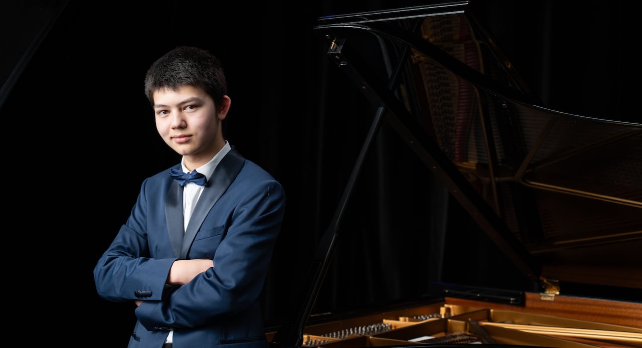 Wearing a dark blue tuxedo, Ryan Huang stands next to a grand piano with the lid open.