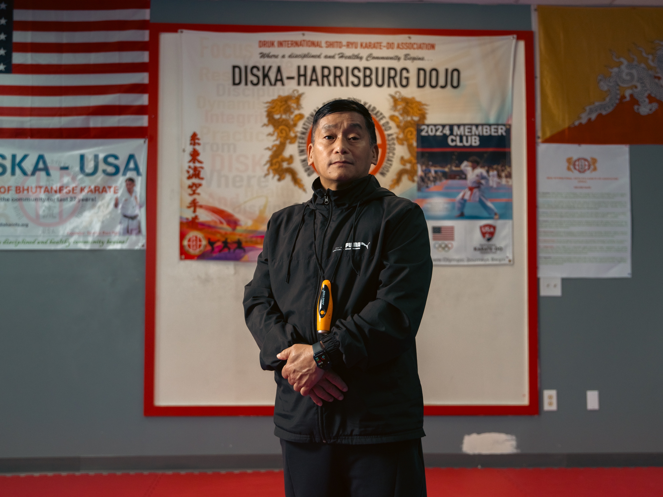 Soke Ran Rai, a martial arts teacher at the DISKA Harrisburg Dojo, began teaching karate in the refugee camps in Nepal before relocating to the United States. He poses for a portrait in the dojo, where the U.S. and Bhutanese flags hang behind him, on April 16, 2025, in Harrisburg, Pa.