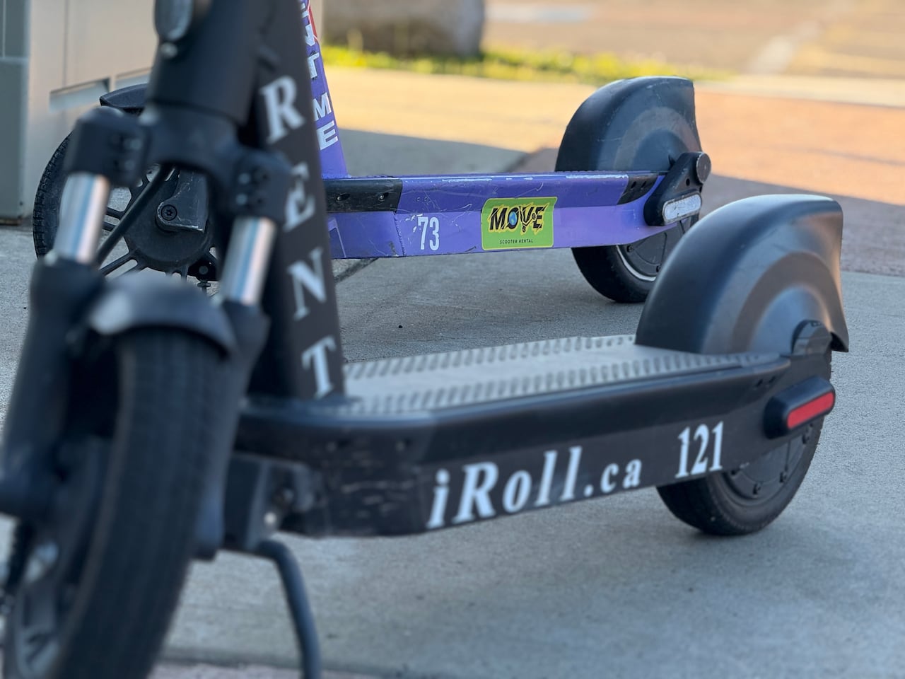 A close-up of two scooters, one in the foreground that's black with the iRoll website and a purple one in the background with the MOVE logo.