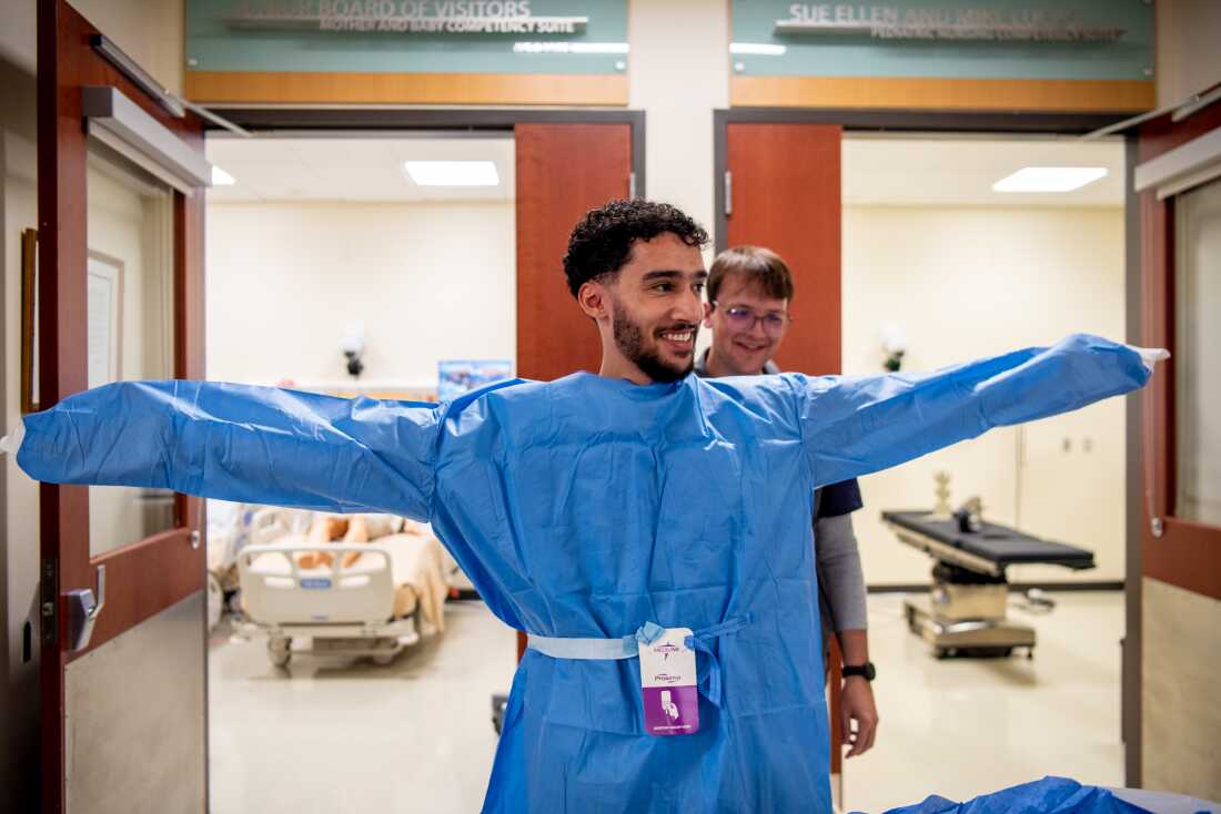 Nursing student Hatim Nasser practices proper installation of personal protective equipment (PPE) with classmates. He is wearing a blue surgical gown over his scrubs with his arms outstretched. He has short, curly dark hair and a short beard.