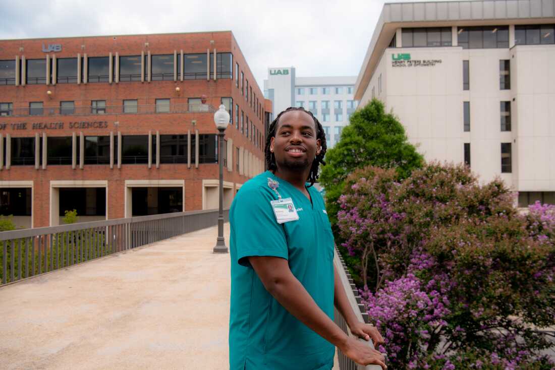 Nursing student Nicholas Seay is wearing teal scrubs and has shoulder length braids. He is standing on a walkway between UAB buildings.