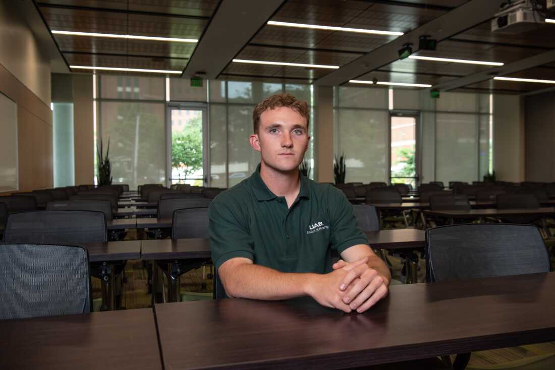 Nursing student Connor Aderholt has short curly hair and is wearing a UAB polo shirt. He is sitting at a table in a classroom.