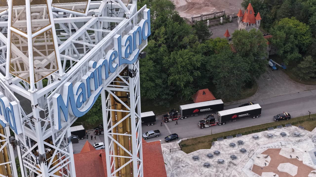 The Marineland scene is seen at the top of an amusement park ride's tower. Four black transport trucks, their flatbeds now covered with black trailers that say "PADDOCK" are on the street below.