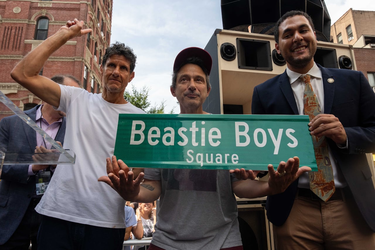 Three men stand in New York holding up a green sign that reads "Beastie Boys Square."