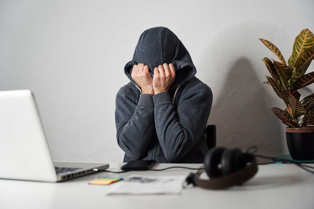 The photo shows a person covering his face with his hands and hoodie at an office desk.