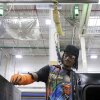 Kwame Crockett is a Black man and he is wearing a black baseball cap, safety glasses and a colorful shirt as he assembles parts of an oven.