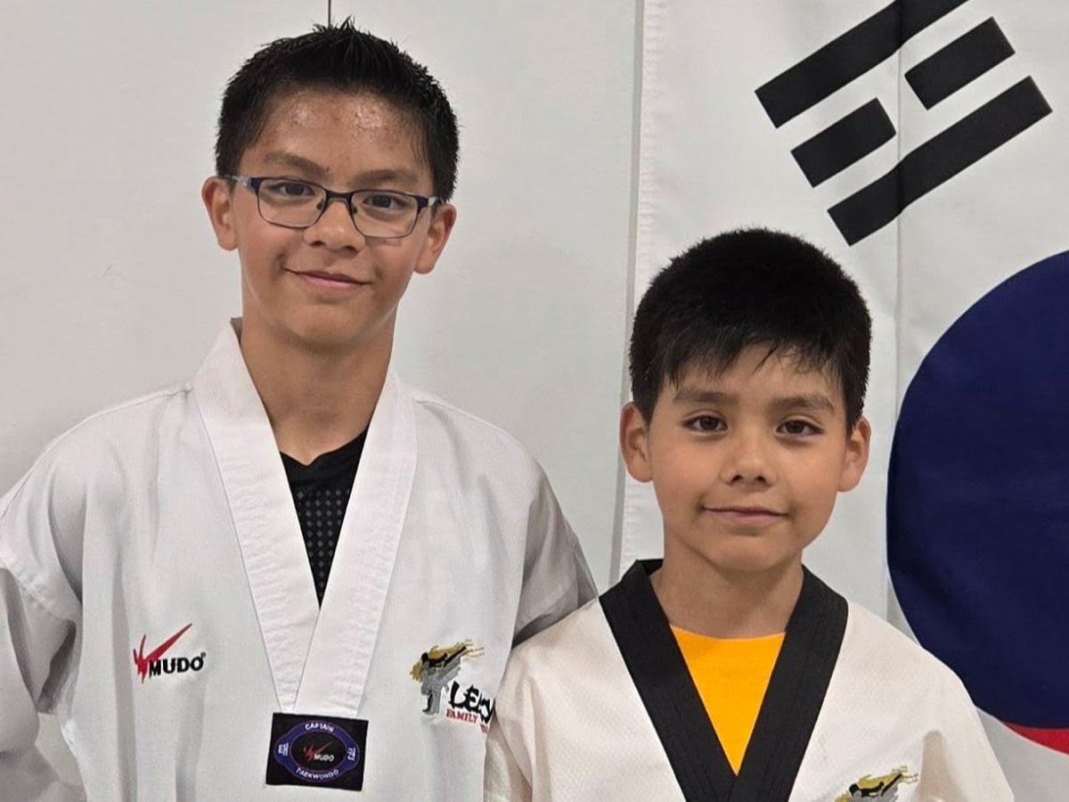 Two boys in white martial arts outfits, with the one on the left wearing glasses and a South Korea flag on a wall behind them