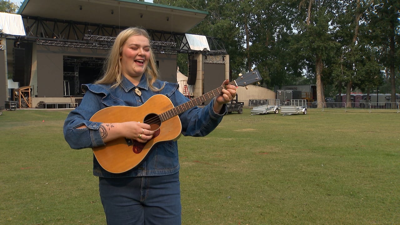 A woman plays a guitar and sings while crews work to set up a stage in the background.