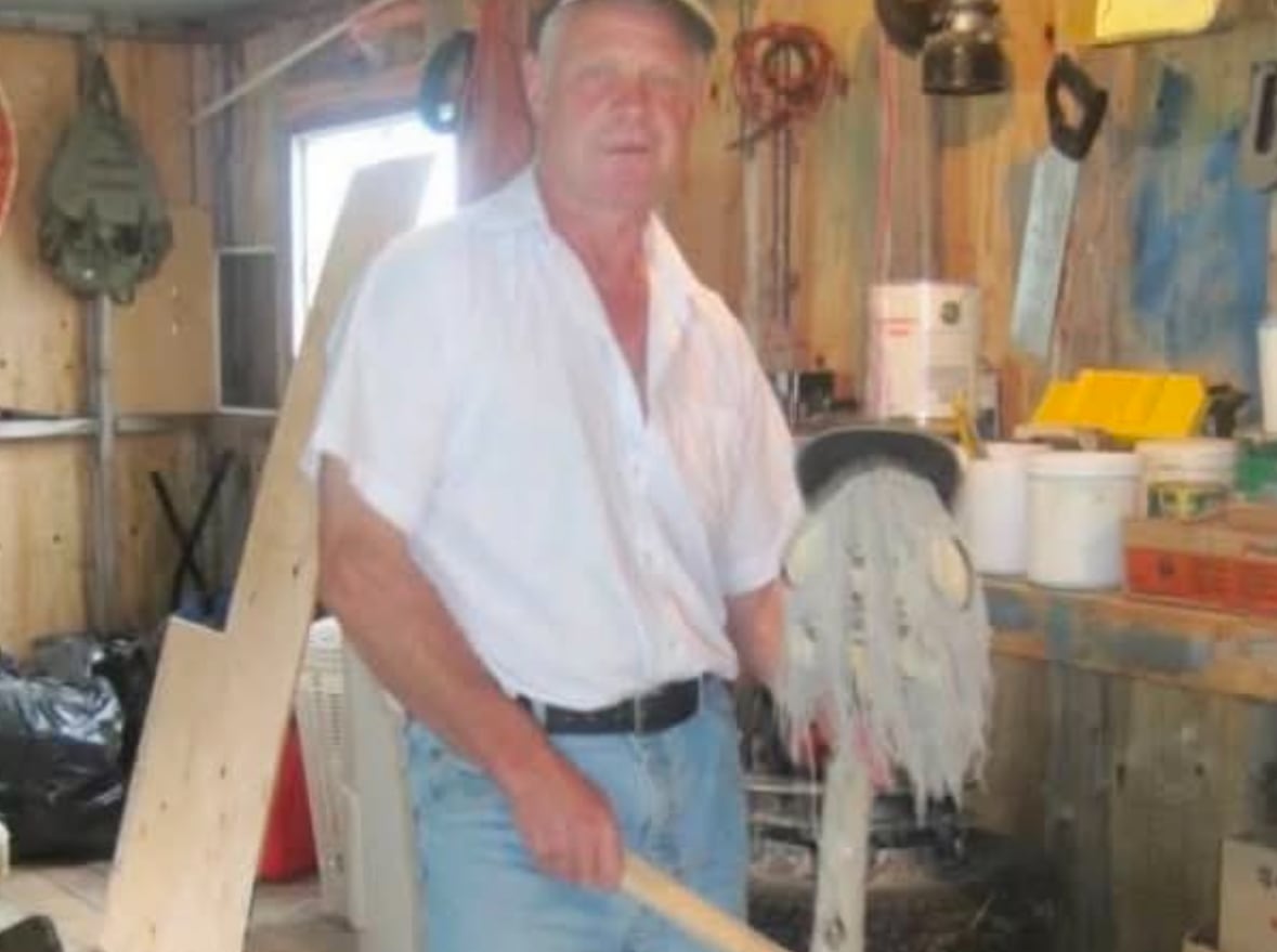 Standing in a shed or workshop, a man in jeans, a white shirt and rubber boots taps an ugly stick.