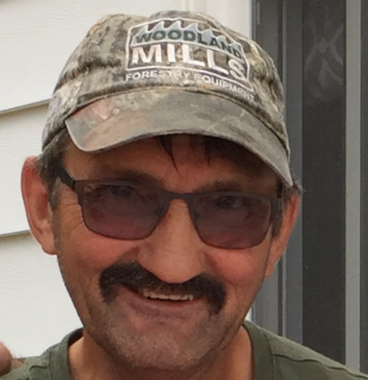 A head-and-shoulders image of a man in sunglasses, a green t-shirt and ballcap. He is smiling.