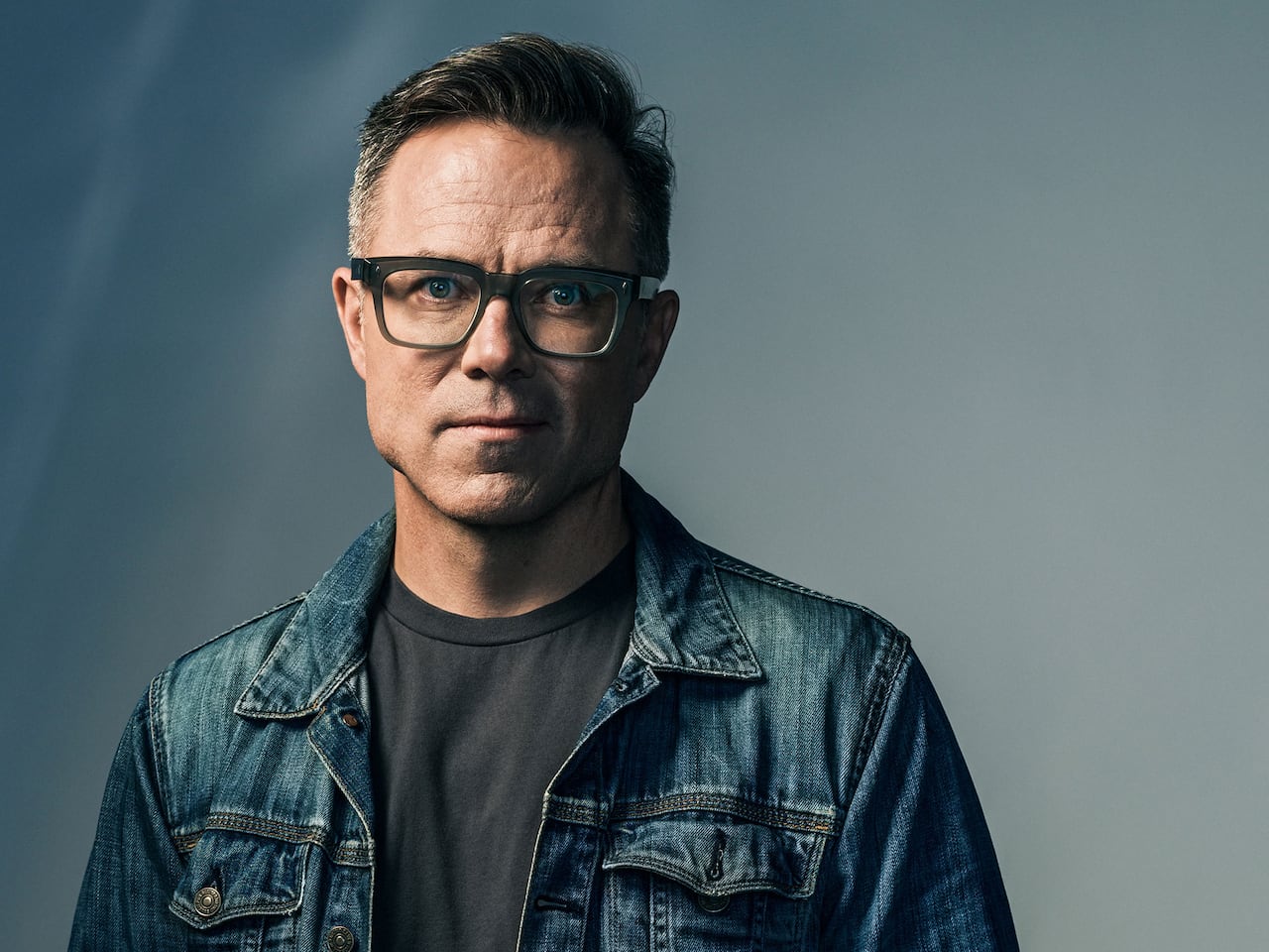 A man wearing glasses and a denim jacket poses for a photo
