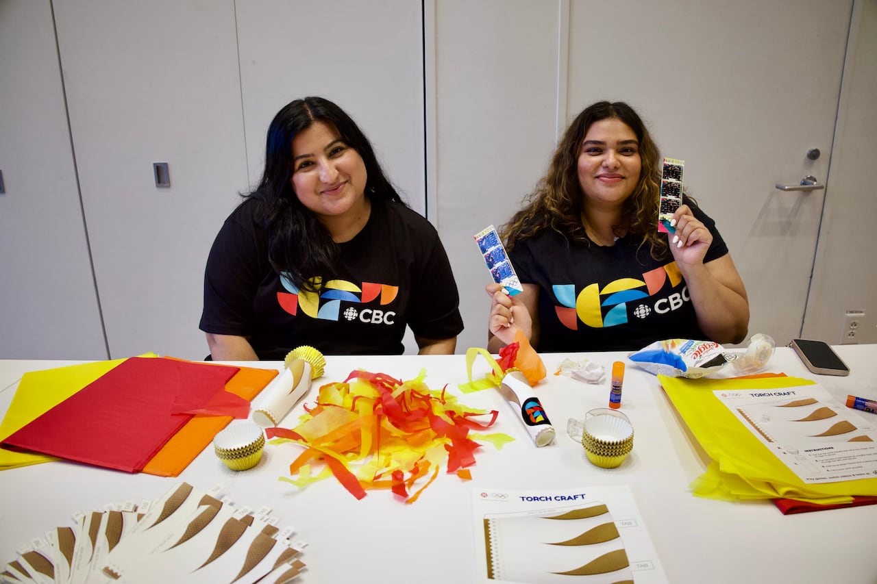 Two women in black CBC t-shirts smile at the camera while sitting at a table filled with colorful crafting materials, including paper, ribbons, and glue.