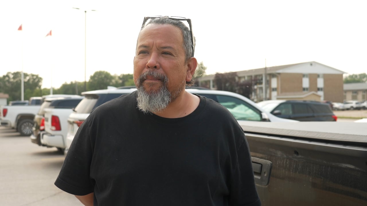 A man with a goatee in a black t-shirt stands in a parking lot.
