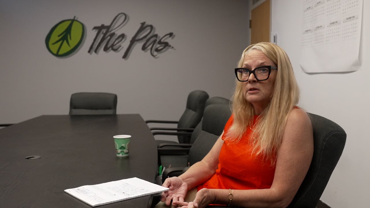 A woman wearing glasses sits at a board room table with the words "The Pas" on a wall in the background.