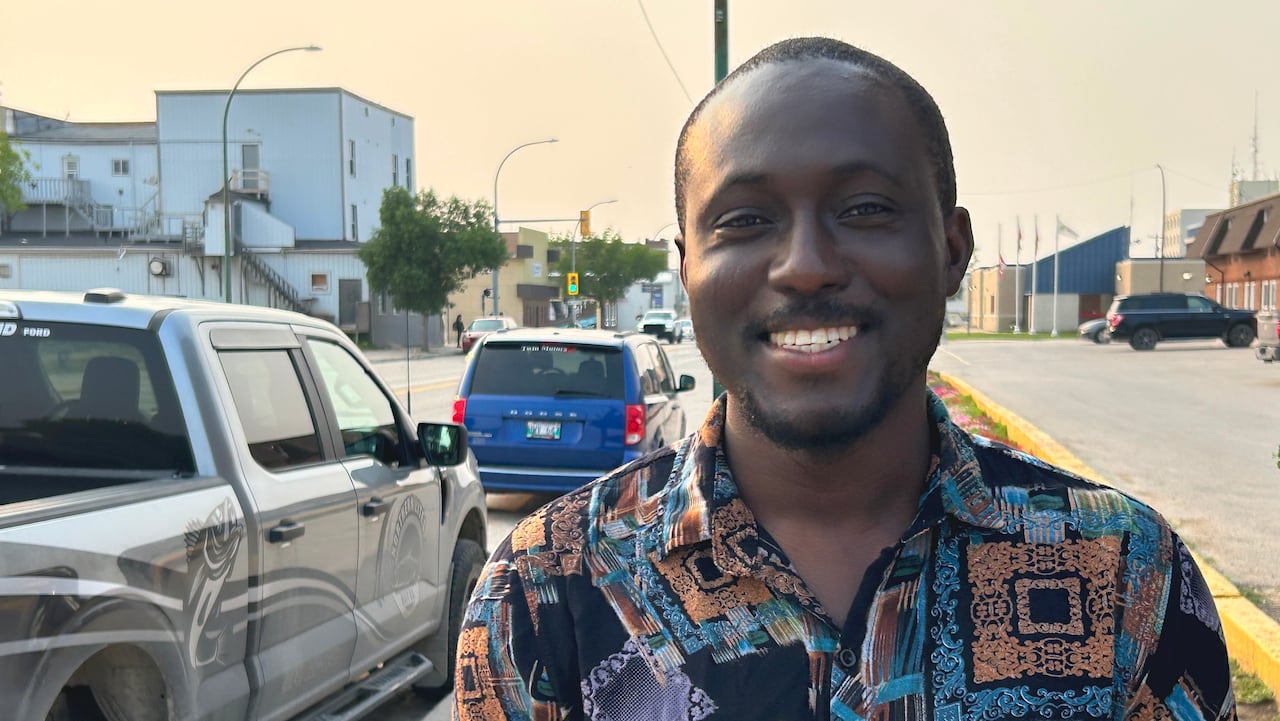 A man in a colourful shirt stands smiling on a sidewalk next to a surface street.
