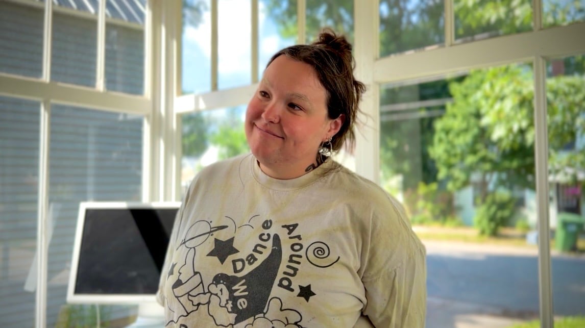 A woman with dark hair in a bun wearing a white graphic shirt stands inside the Mudroom Gallery porch on a sunny day.