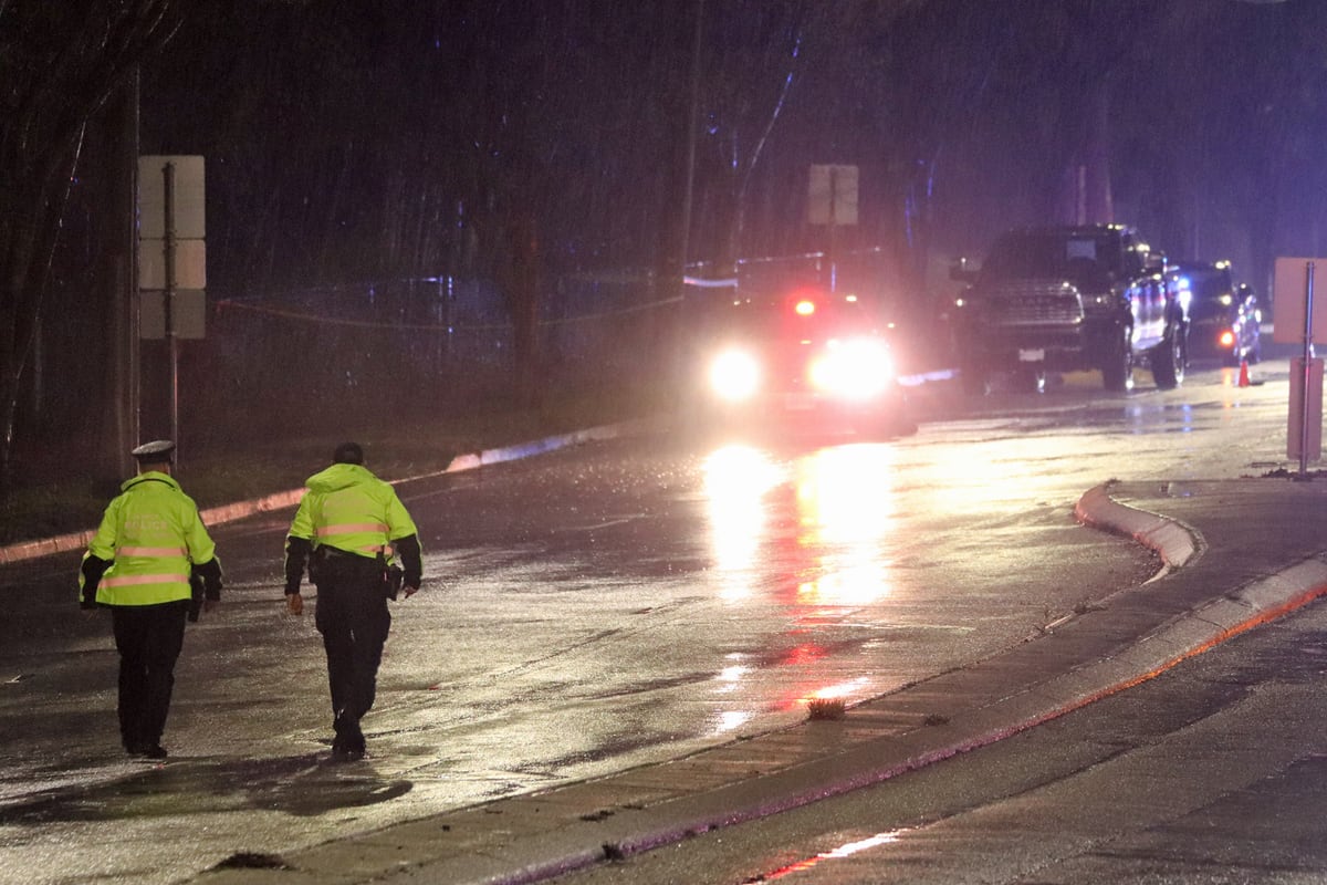Police officers walk on a rainy street with police cars in the distance.