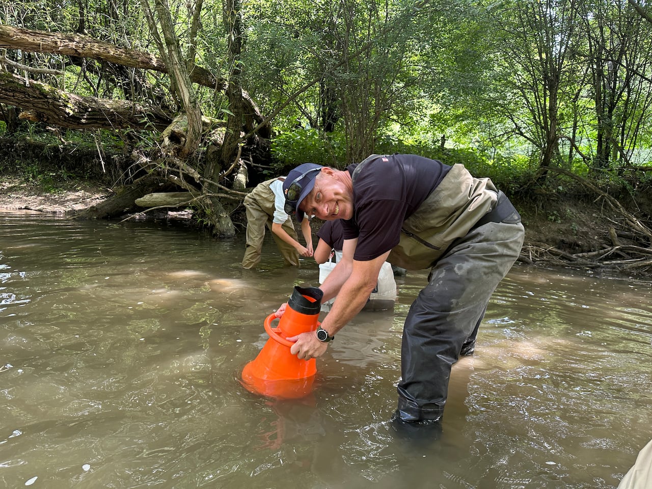 A man wearing green chest waders holds an orange cone in the water.