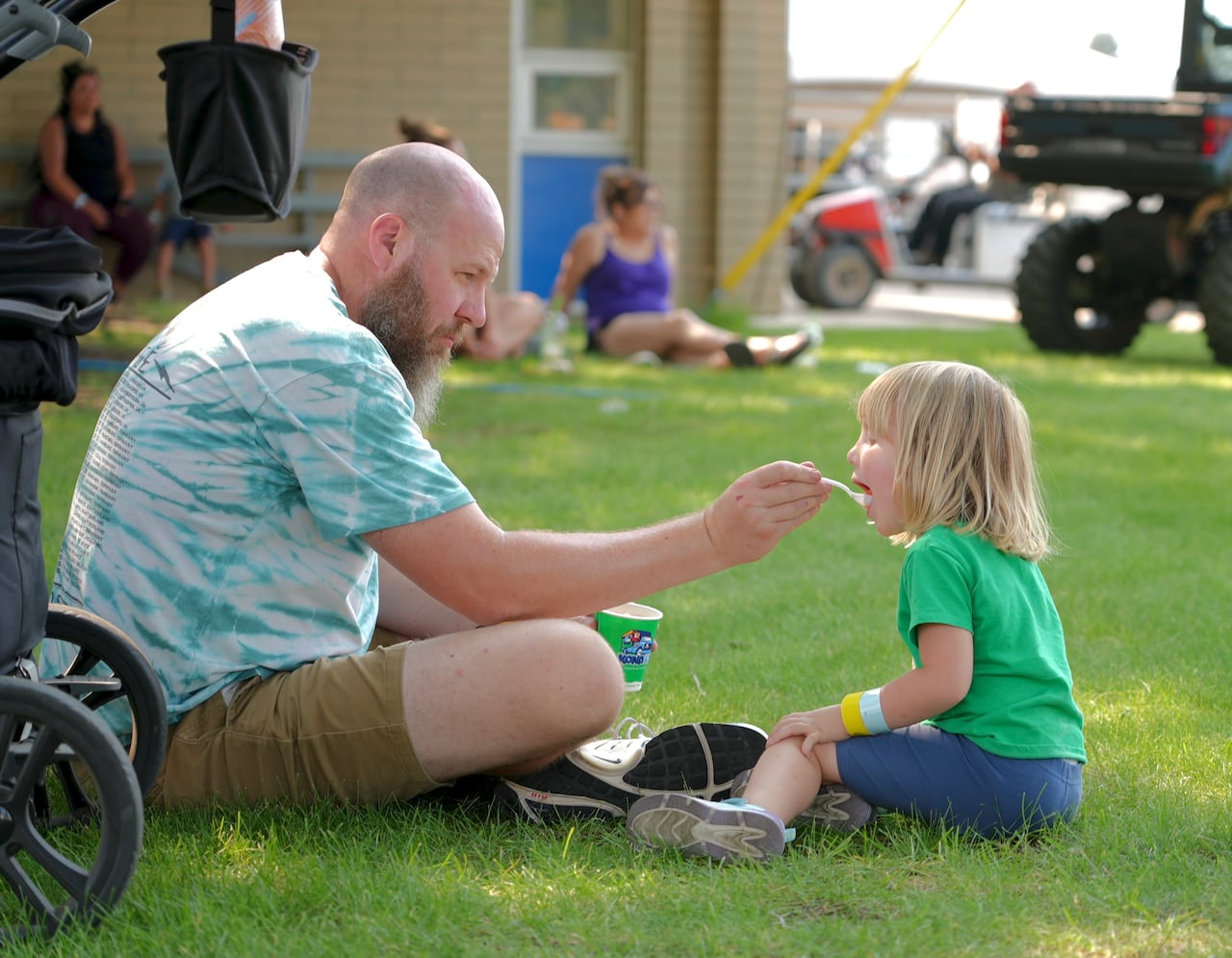 A large man with a broad shoulders holds out a spoon of shaved ice for his toddler as they sit crosslegged on the grass.